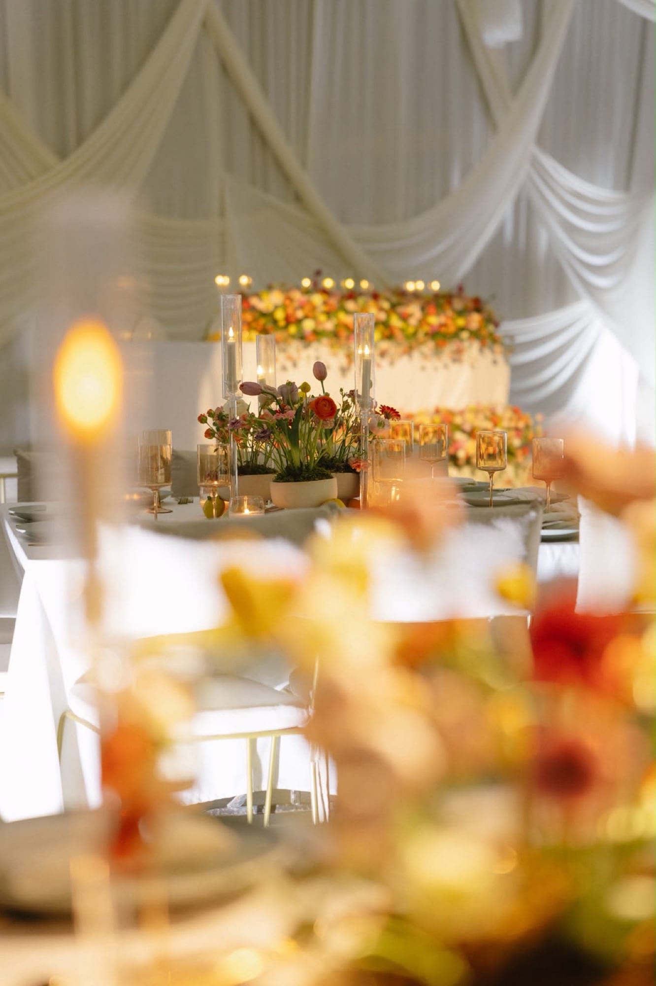 Mesa con velas altas y flores cálidas bajo un techo drapeado en blanco
